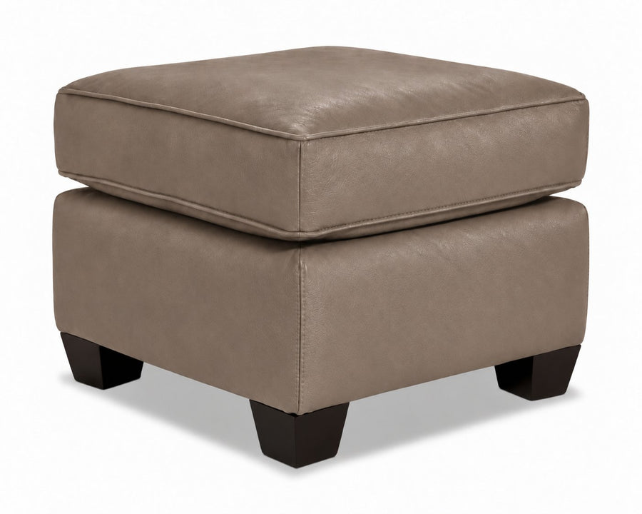 City Leather Ottoman - Stone in a three-quarter view with a thick piped cushion, leather upholstery and dark wood legs
