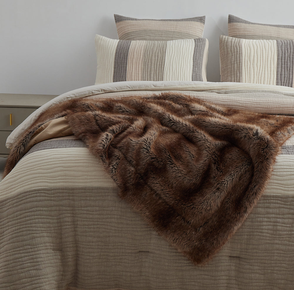 The Great Indoors light brown faux fur throw draped across a bed with neutral striped bedding.